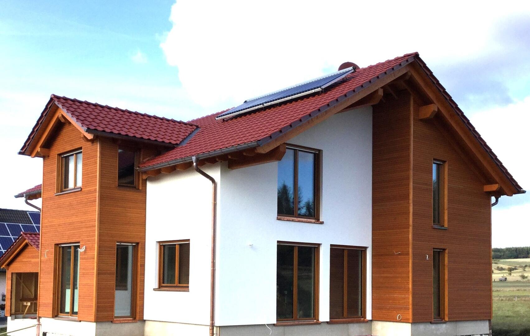 Ein modernes zweistöckiges Haus mit rotem Ziegeldach, großen Fenstern und einer Mischung aus weißen und holzgetäfelten Außenwänden. Auf dem Dach sind Solarmodule installiert, und das Haus ist von offener Landschaft umgeben.