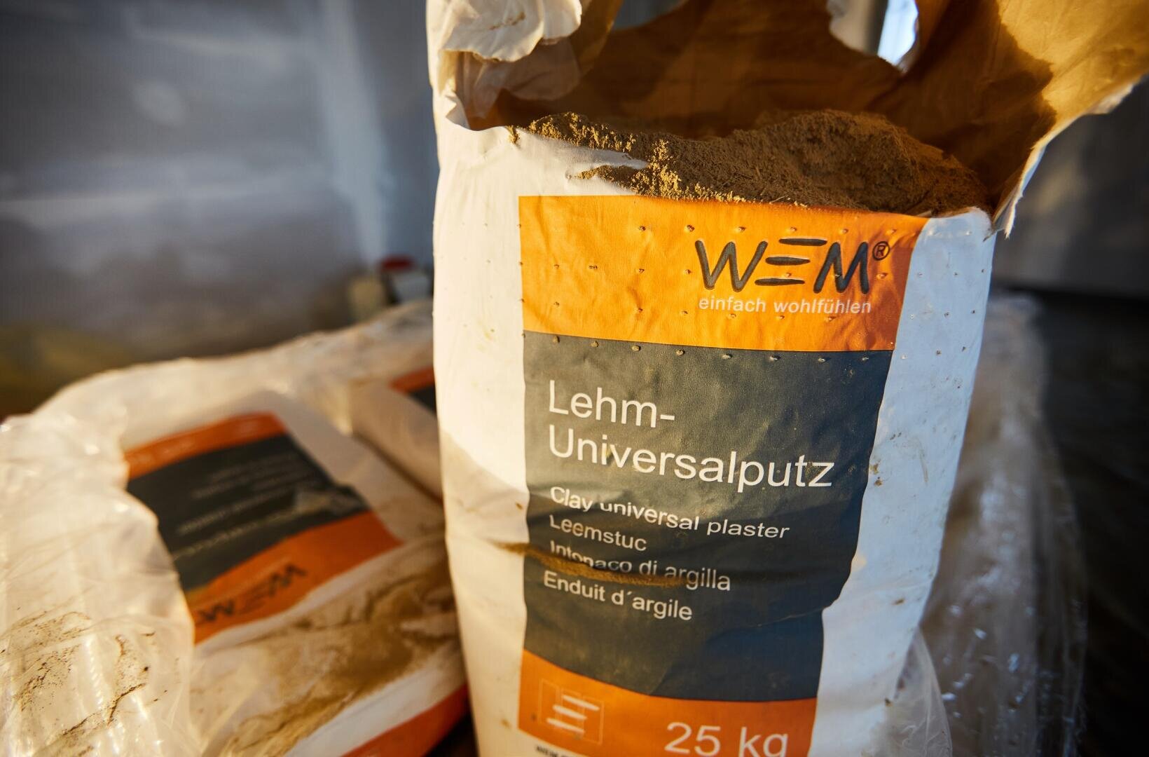 Nahaufnahme eines 25-kg-Sacks WEM Lehm-Universalputz. Der geöffnete Sack gibt den Blick auf das Pulver frei. Der Sack ist orange-grau beschriftet und liegt auf ähnlichen Säcken.