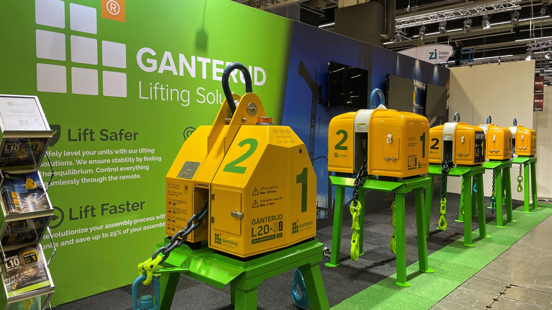 Eine Reihe gelber industrieller Hebevorrichtungen auf grünen Ständern auf einem Messestand von Ganterud Lifting Solutions, mit Informationsschildern und Broschüren im Hintergrund.