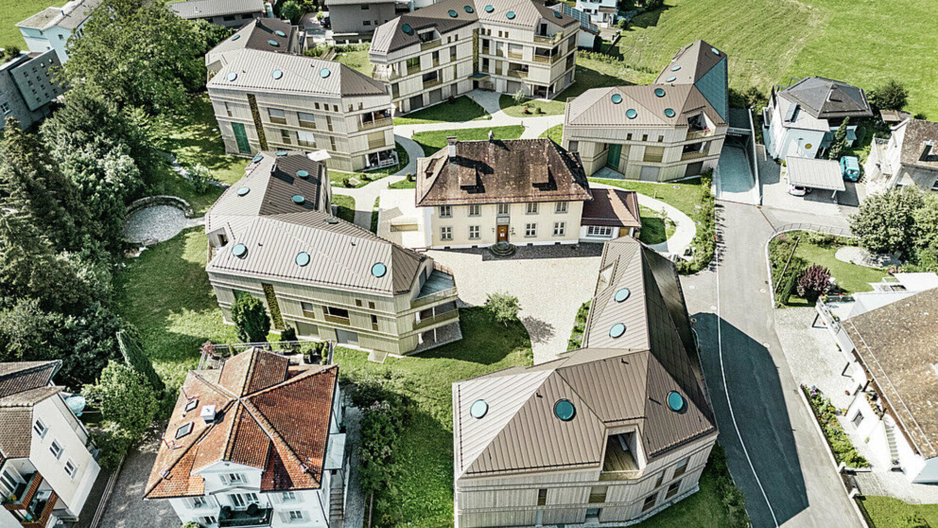 Luftaufnahme moderner Wohngebäude, die um ein zentrales, älteres Haus mit Innenhof angeordnet sind; grüne Rasenflächen und Bäume umgeben die Gebäude, in der Nähe befinden sich eine Straße und weitere Häuser.
