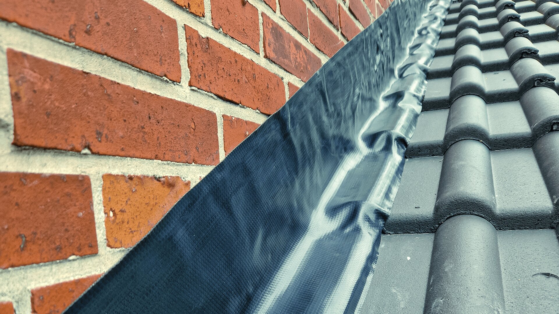 Nahaufnahme einer Ziegelwand, die auf ein Ziegeldach trifft. An der Fuge ist eine schwarze Schutzleiste angebracht, um Wassereintritt zu verhindern. Die Schutzleiste ist sorgfältig befestigt, und die Dachziegel sind dunkel und gleichmäßig angeordnet.