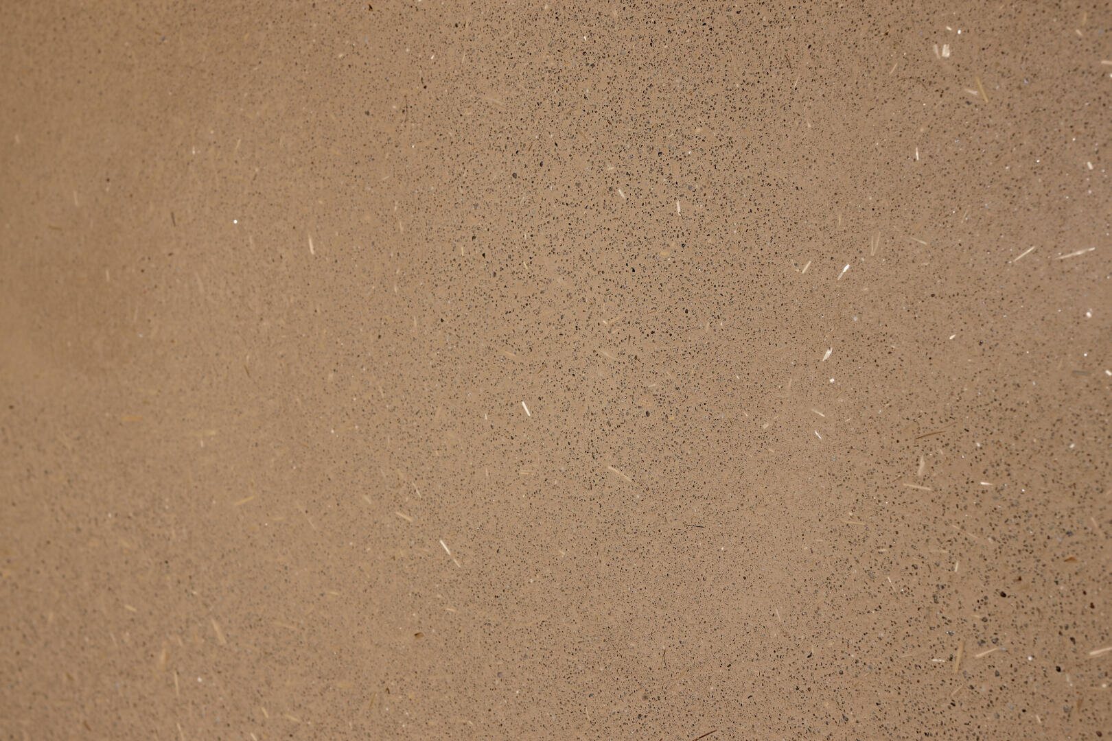Nahaufnahme einer strukturierten beigen Oberfläche mit kleinen schwarzen Flecken und hellen Streifen. Die Textur wirkt rau und ähnelt möglicherweise Beton, Stein oder Gips.