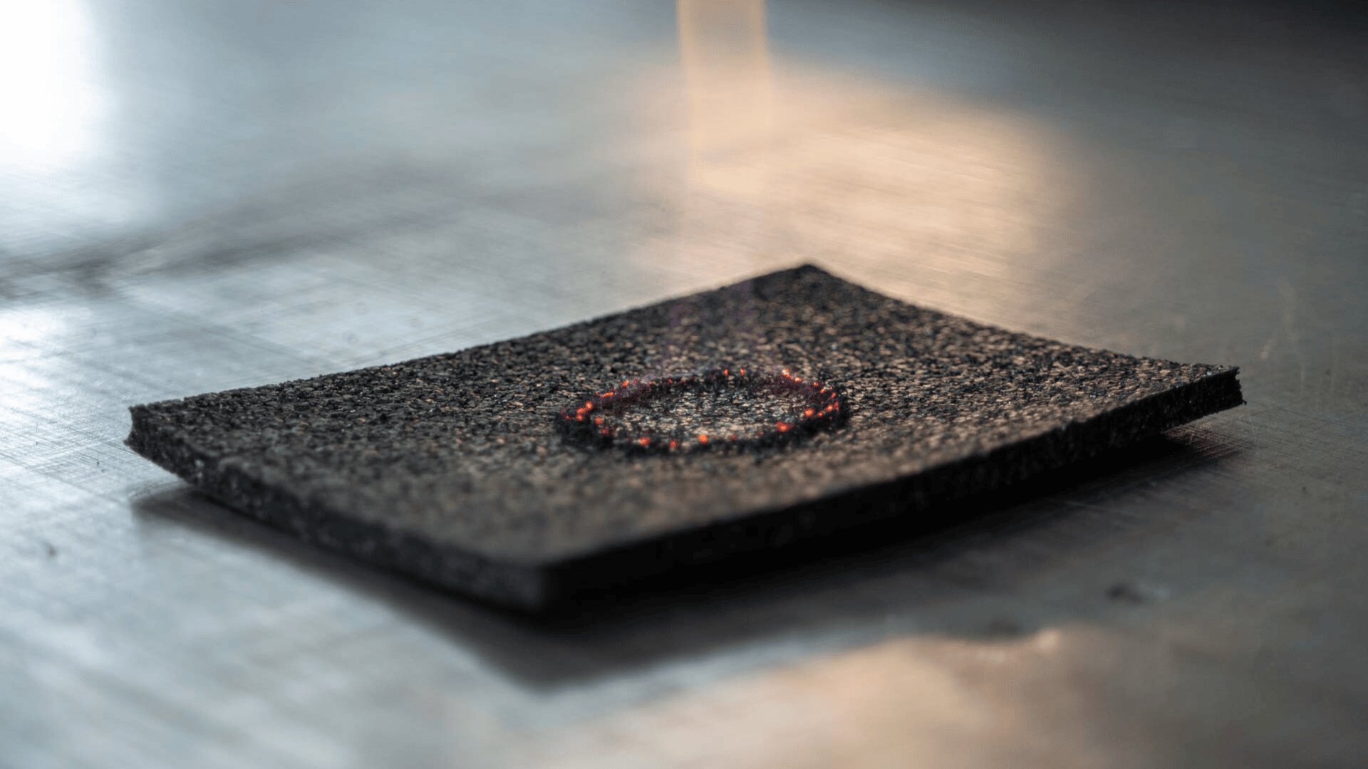 Nahaufnahme eines schwarzen, strukturierten Materials mit einem kreisförmigen Fleck aus Glut und Rauch in der Mitte, der auf einer glatten, metallischen Oberfläche liegt.