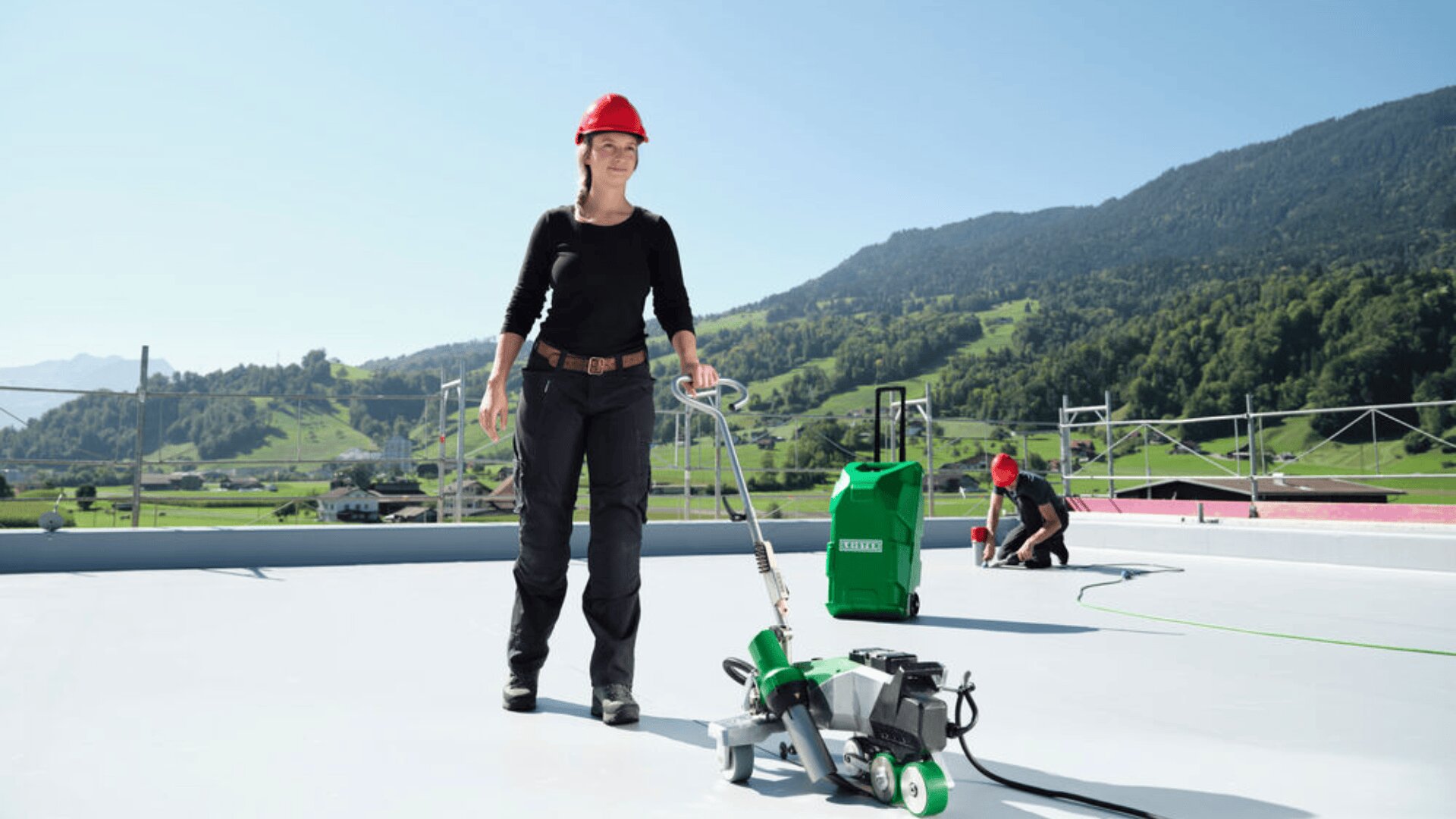 A person in a red hard hat operates roofing equipment on a flat roof, with mountains and greenery in the background. Another person in a hard hat works in the distance. The weather is clear and sunny.