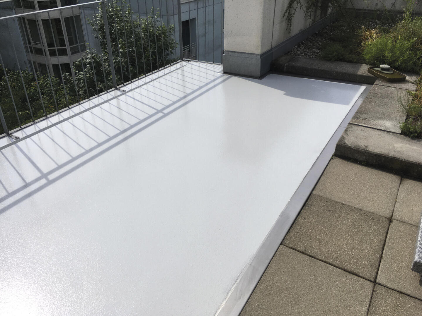 Eine Dachterrasse mit glänzender, neu beschichteter weißer Oberfläche neben beigen quadratischen Terrassenfliesen, Metallgeländern und Pflanzkübeln mit Grünzeug im Hintergrund.