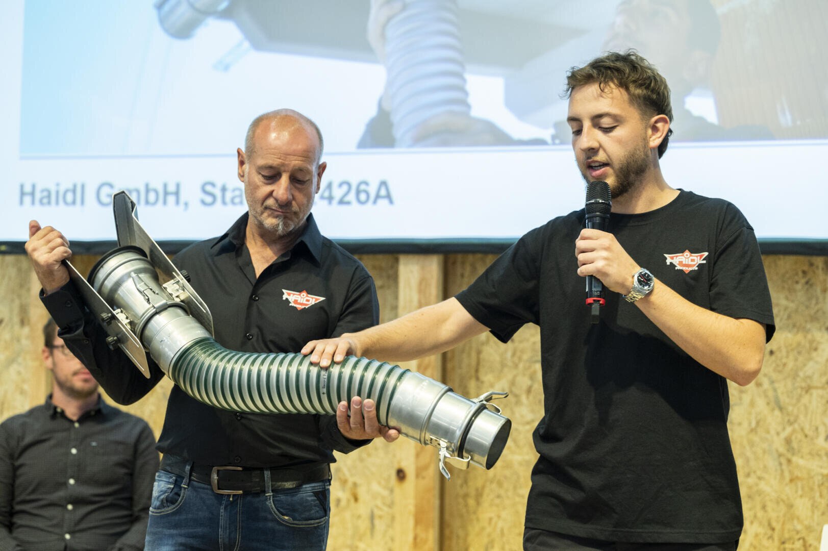 Zwei Männer stehen nebeneinander. Einer hält einen großen Industrieschlauch mit Metallbeschlägen, während der andere in ein Mikrofon spricht und auf den Schlauch deutet. Im Hintergrund ist ein Bildschirm mit Textausschnitten zu sehen.
