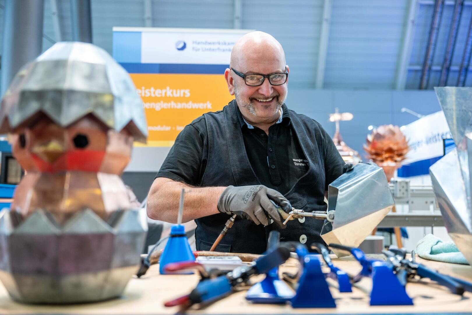 Ein lächelnder Mann mit Brille und Handschuhen arbeitet mit einem Lötwerkzeug an einer Werkbank. Im Vordergrund stehen Metallskulpturen, darunter ein Küken, das aus einem Ei schlüpft, und verschiedene Werkzeuge.