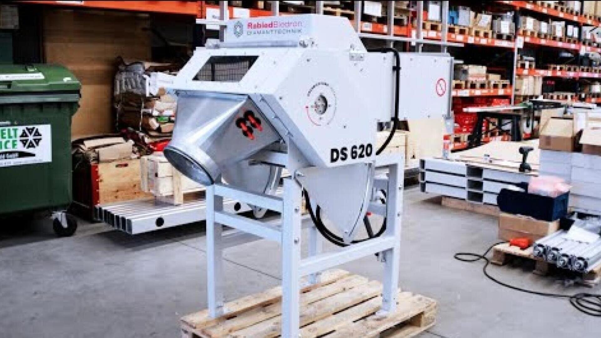Industriemaschine mit der Bezeichnung DS 620 und trichterförmigem Ausgang, montiert auf einer Holzpalette in einem Lager, umgeben von Regalen mit verschiedenen Kisten und Geräten.