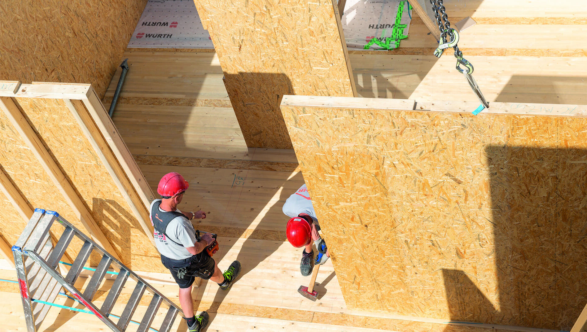 Zwei Bauarbeiter mit roten Helmen arbeiten in einem Holzgebäude. Eine Leiter steht daneben, und Bauwerkzeuge sind sichtbar. Sonnenlicht wirft Schatten auf die Holzwände und den Boden.