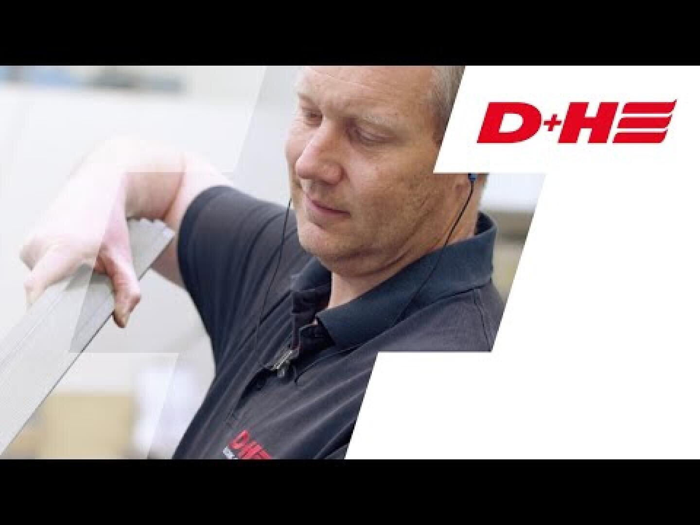 Ein Mann in einem schwarzen Poloshirt mit rotem D+H-Logo untersucht einen langen Metallgegenstand. Der Hintergrund ist überwiegend weiß, mit einem großen roten D+H-Logo auf der rechten Seite.