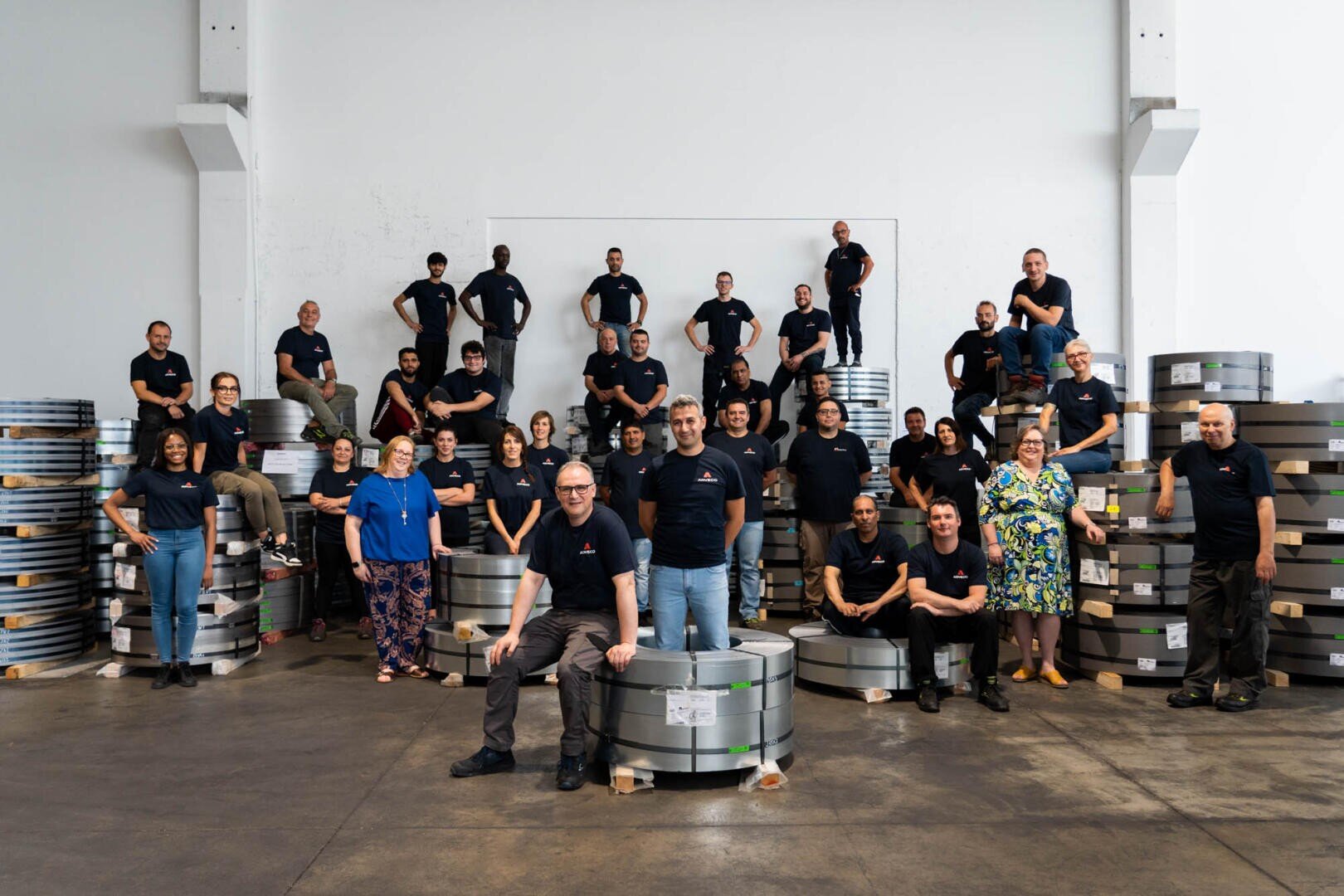 A group of about 35 people in matching dark shirts pose smiling in a warehouse with stacks of metal coils around them. Some stand, others sit or kneel against the backdrop of a white wall.