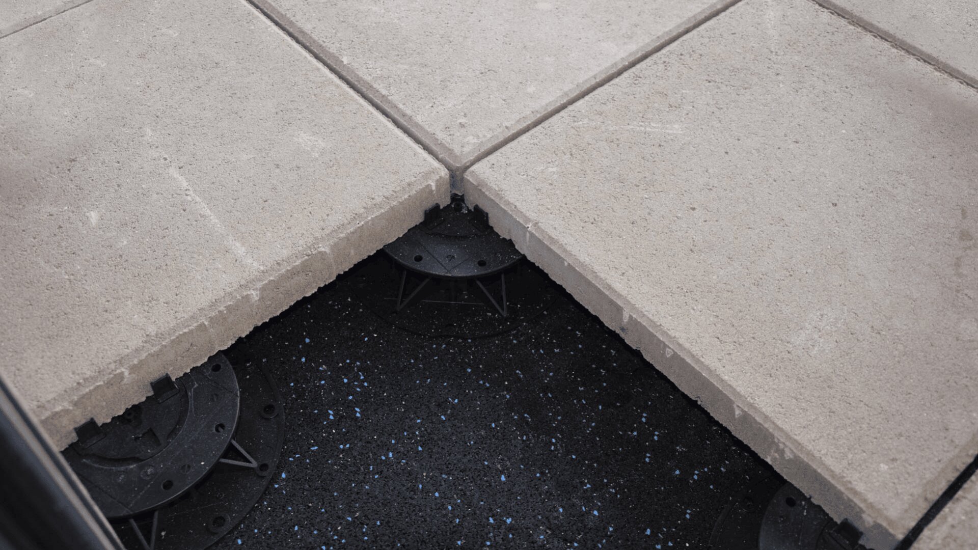 Nahaufnahme von großen Betonpflastersteinen mit einer angehobenen Fliese, die schwarze Sockelstützen und eine gesprenkelte dunkle Oberfläche darunter erkennen lässt.