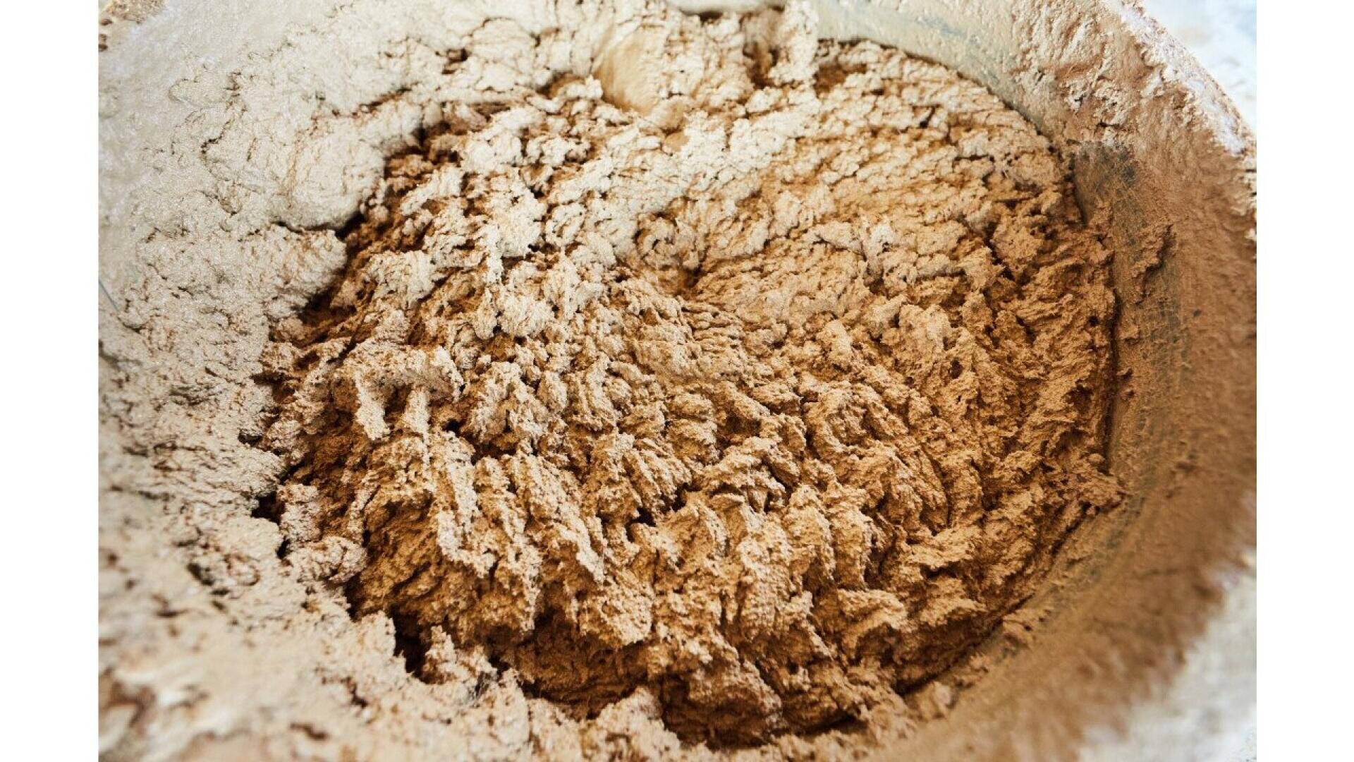 A close-up of a mixing bowl filled with dry, light brown powder, likely flour or a baking mix, showing a rough, uneven surface with clumps and textured patterns.