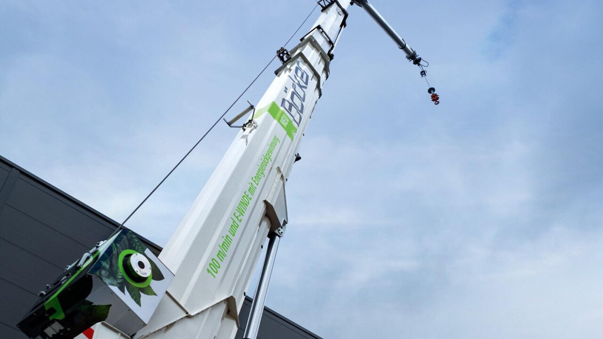 A large white construction crane extends upward against a cloudy sky, with green and black markings and text visible on its side. The cranes arm has a hook at the end, and part of a building is visible on the left.