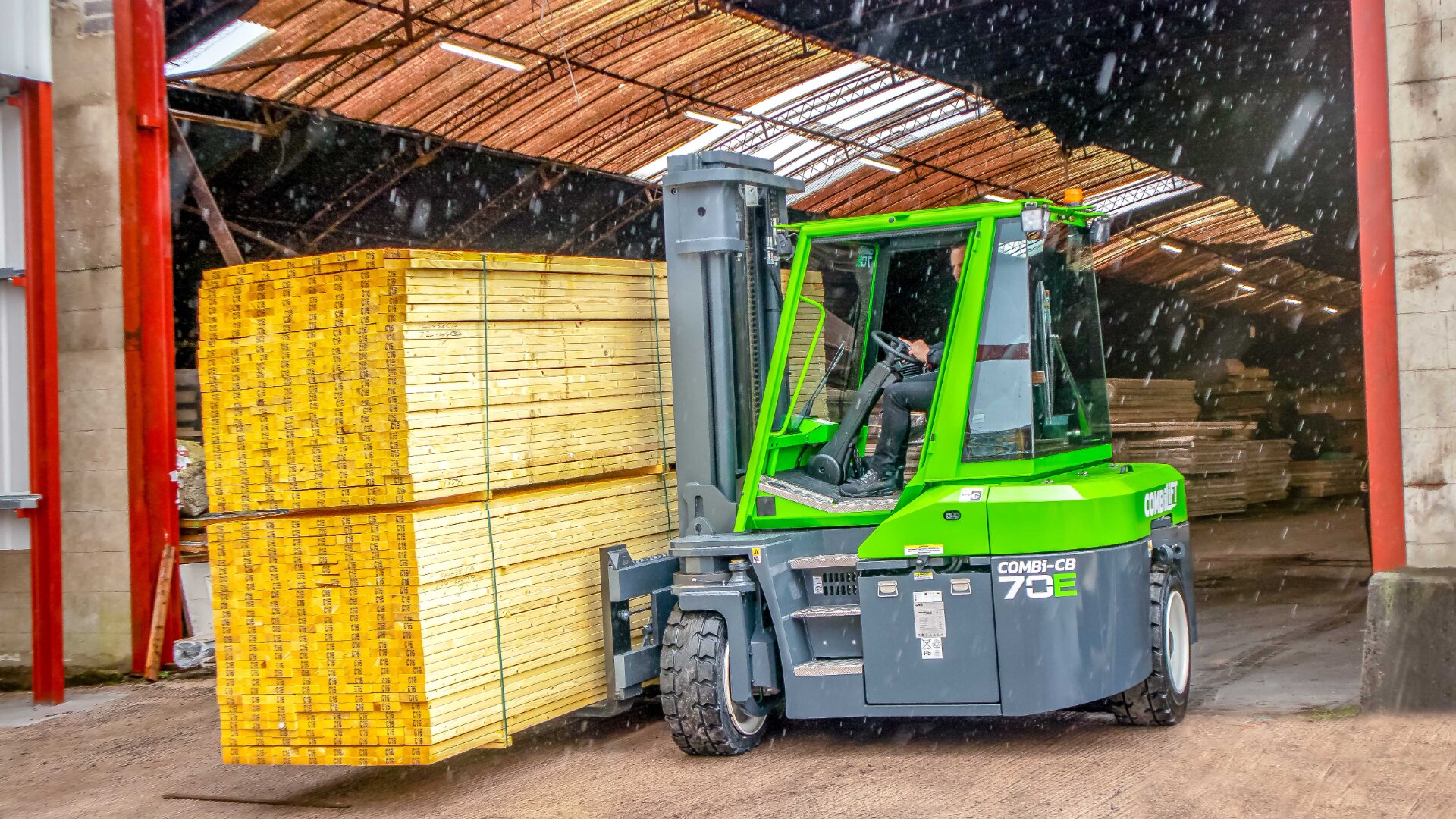 Ein grüner Gabelstapler transportiert einen großen Stapel gelber Holzbretter aus einer Lagerhalle. In der Luft rund um das Fahrzeug sind Schnee- oder Sägemehlpartikel zu sehen.