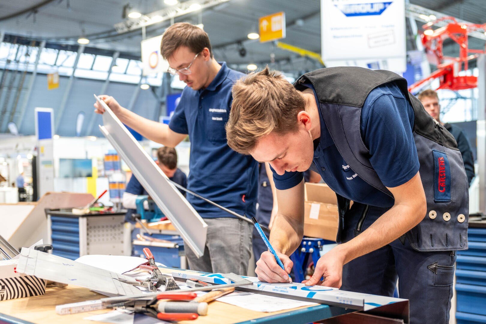 Zwei junge Männer in blauen Uniformen arbeiten an einem Tisch mit Werkzeugen und elektrischen Bauteilen in einer hellen, industriellen Umgebung. Der eine schreibt auf einem Papier, während der andere eine Schalttafel prüft. Beide scheinen konzentriert und zielstrebig an ihren Aufgaben zu arbeiten.