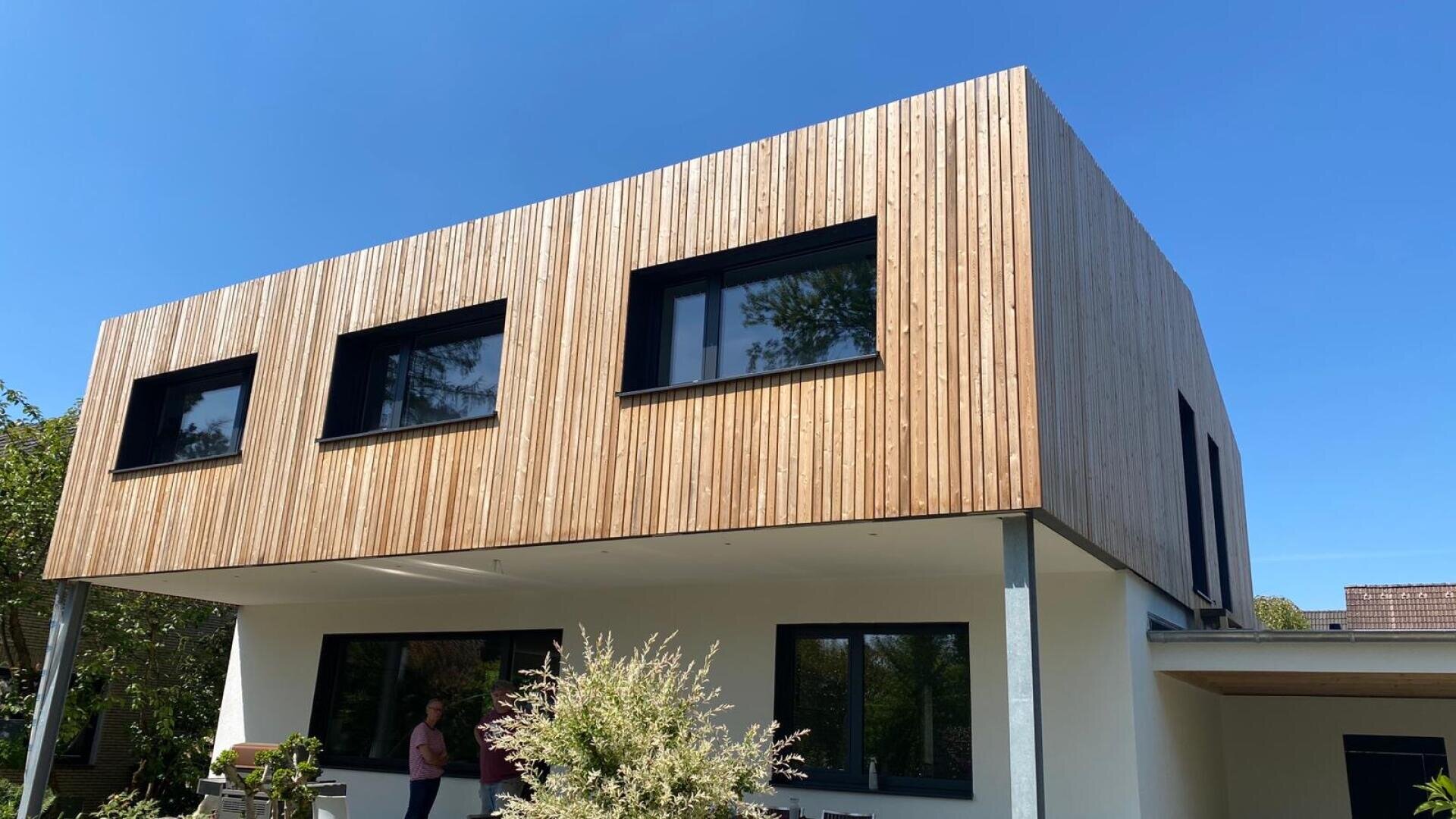 Ein modernes zweistöckiges Haus mit einer Holzfassade im oberen Bereich, rechteckigen Fenstern mit schwarzen Rahmen und einem weißen Untergeschoss. Eine Person steht in der Nähe des Eingangs, und das Haus ist von Grün umgeben, der Himmel ist klar und blau.