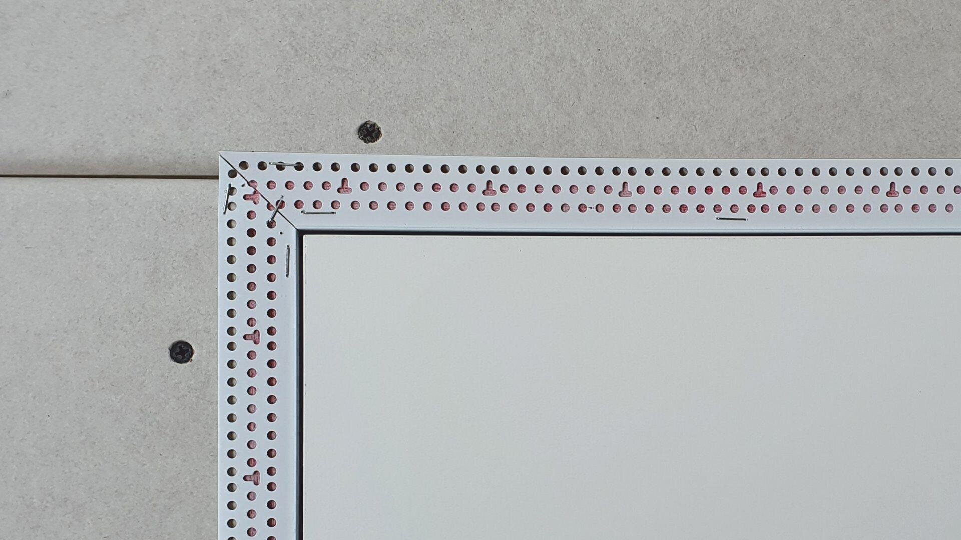 A close-up of a drywall panel with a metal corner bead installed around a rectangular opening, fastened with visible screws. The drywall is light gray, and the metal bead has perforations and red markings.