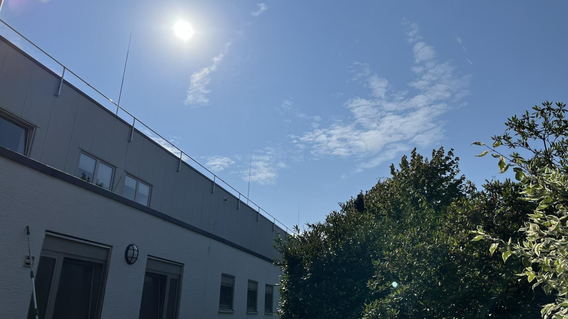 Eine helle Sonne scheint in einem klaren blauen Himmel über einem modernen weißen Gebäude, dessen Vordergrund von Bäumen und grünem Laub eingerahmt wird. Wispy Wolken sind über den Himmel verstreut.