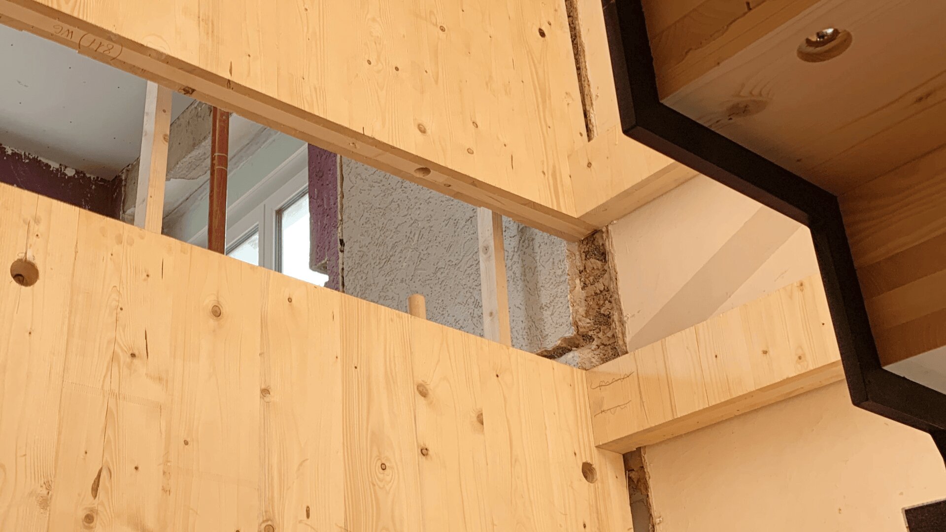 Sichtbare Holzbalken und -paneele in einem im Bau befindlichen Gebäude, im Hintergrund sind eine unfertige Wand und ein Fenster zu sehen.