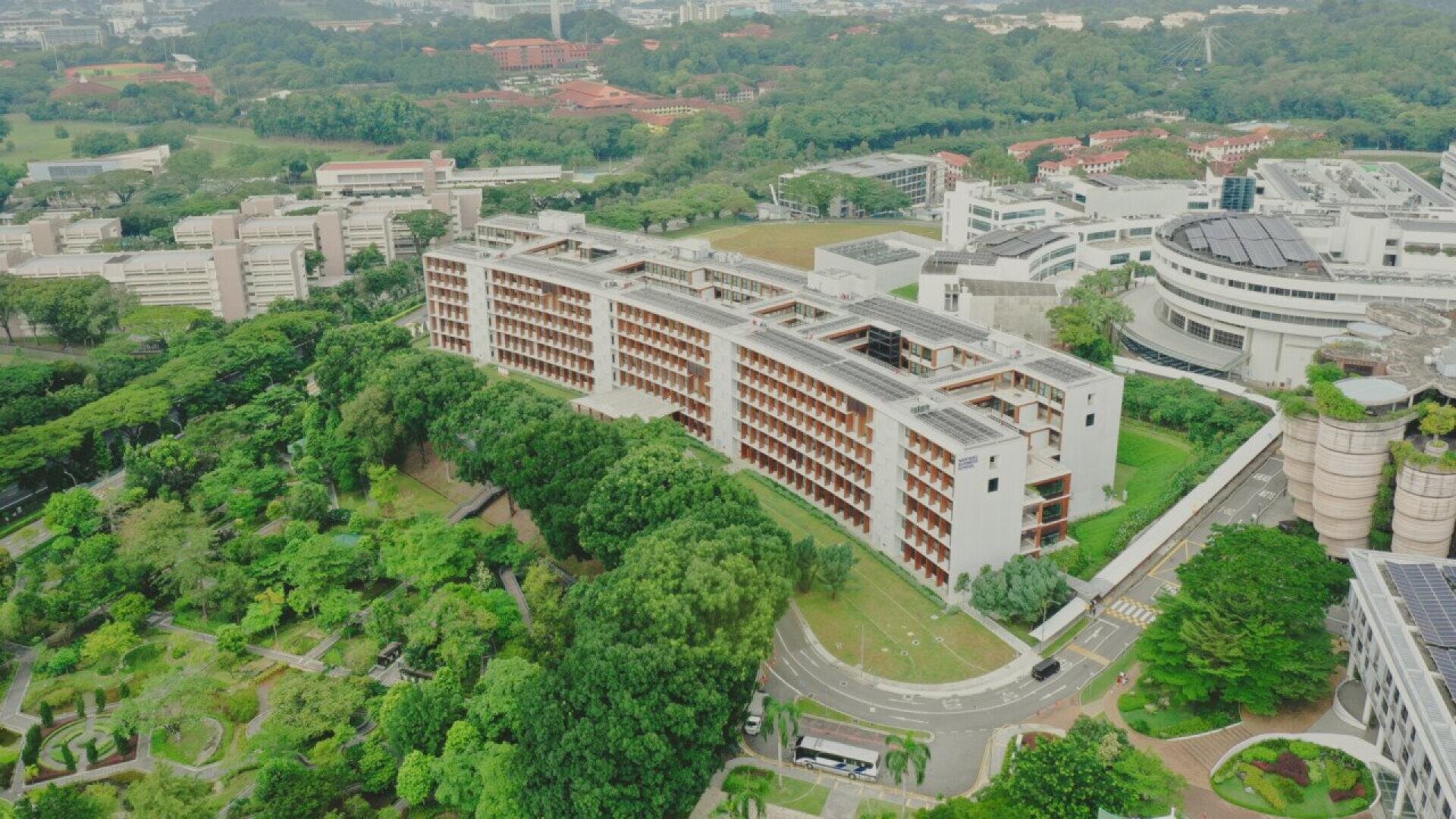 Aerial view of a large modern university campus surrounded by lush greenery, with several buildings, winding roads, and forested areas visible.