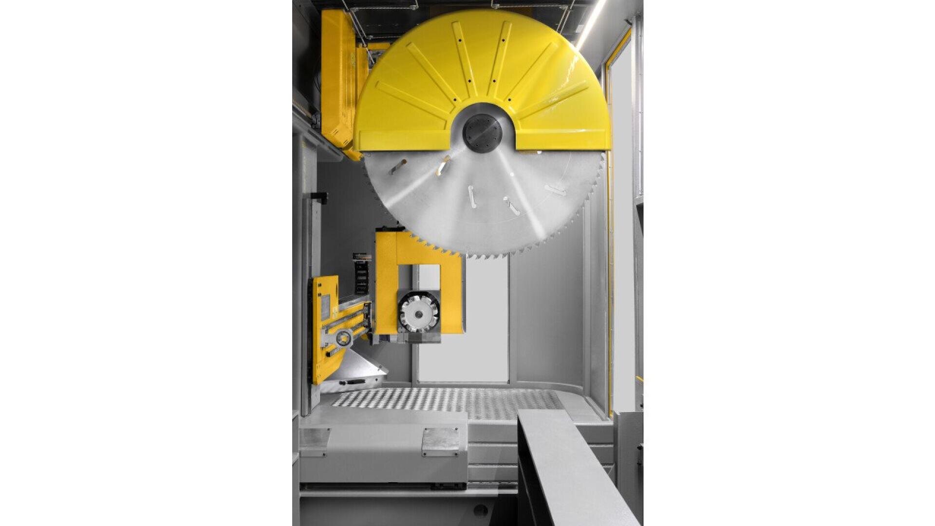 A large, industrial circular saw with a bright yellow safety cover is mounted vertically inside a cutting machine. The interior features metallic components and a gridded work surface below the blade.