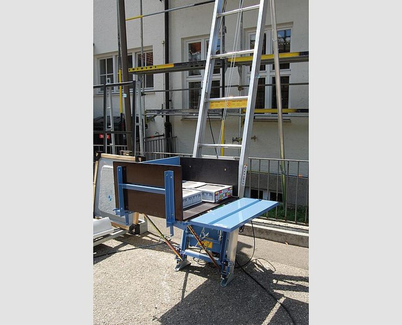A building materials elevator with a blue base and platform is set up outdoors next to a building, with a ladder running vertically above it. Boxes are loaded onto the platform for lifting.