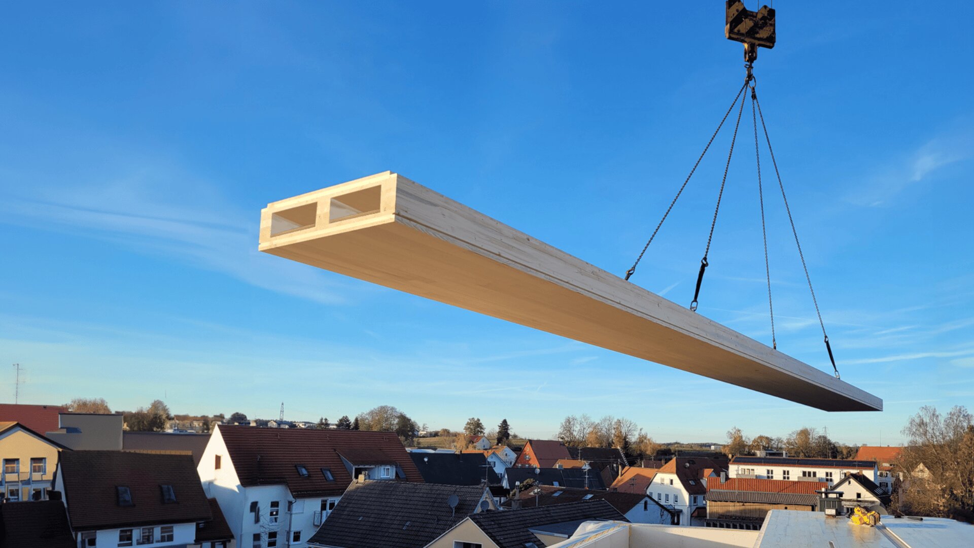 Unter einem klaren, blauen Himmel wird ein großer vorgefertigter Holzbalken von einem Kran über die Dächer eines Wohngebiets gehoben.