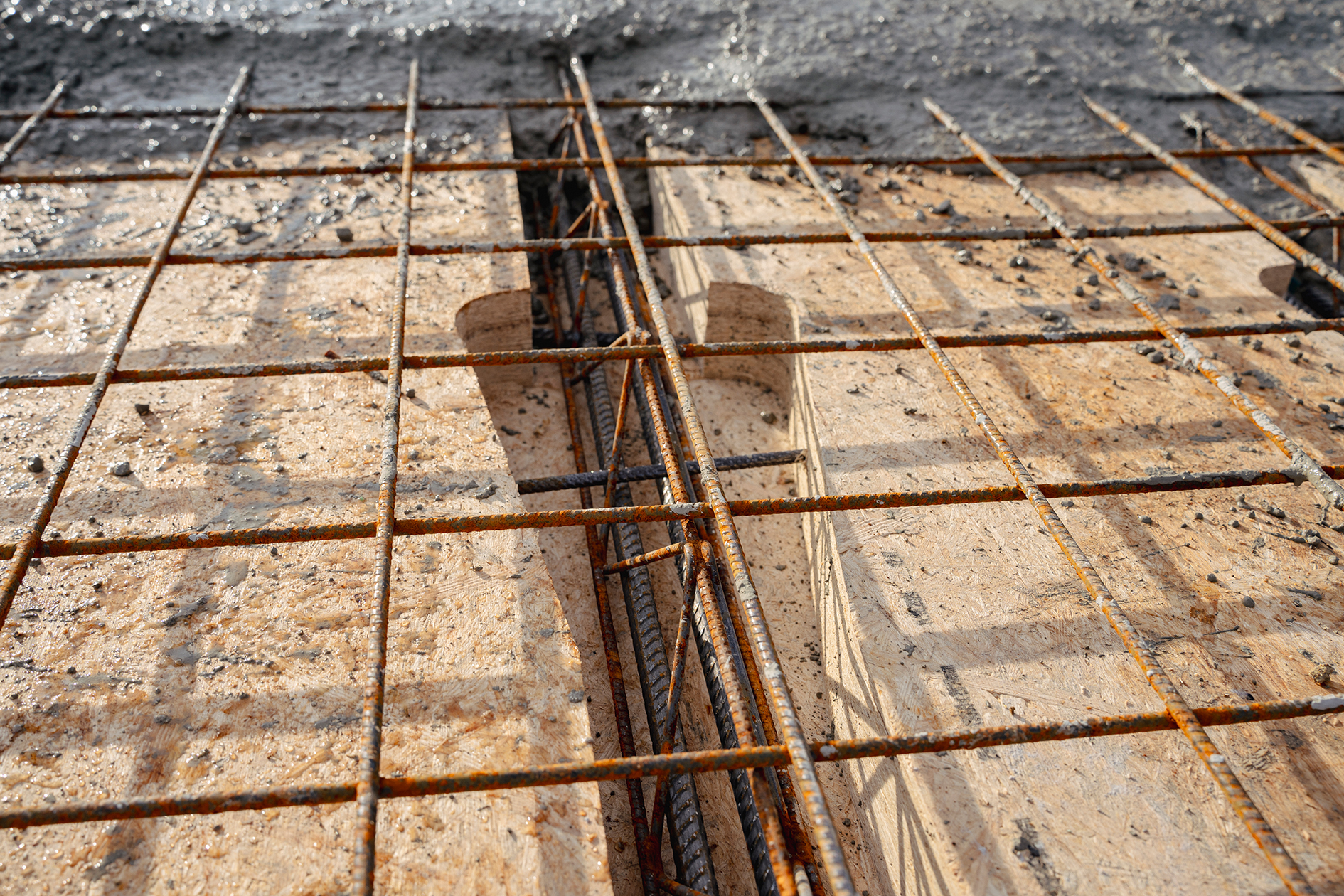 Rusty steel reinforcement bars (rebar) forming a grid pattern on a wooden formwork, prepared for concrete pouring at a construction site. Wet concrete and debris are visible in the background.