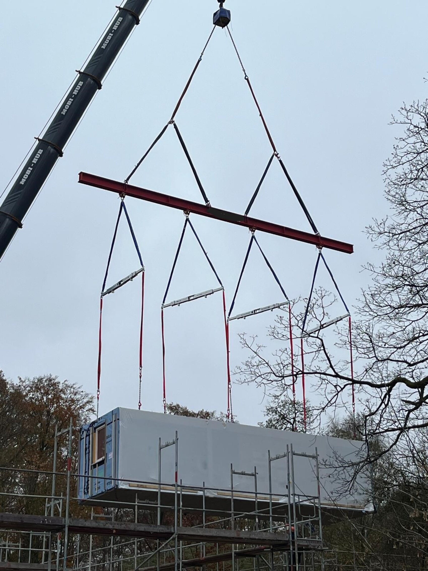 Ein Kran hebt ein großes rechteckiges Fertigbaumodul mit roten Gurten über ein Gerüst und Bäume, um es auf einer Baustelle unter einem bewölkten Himmel zu platzieren.