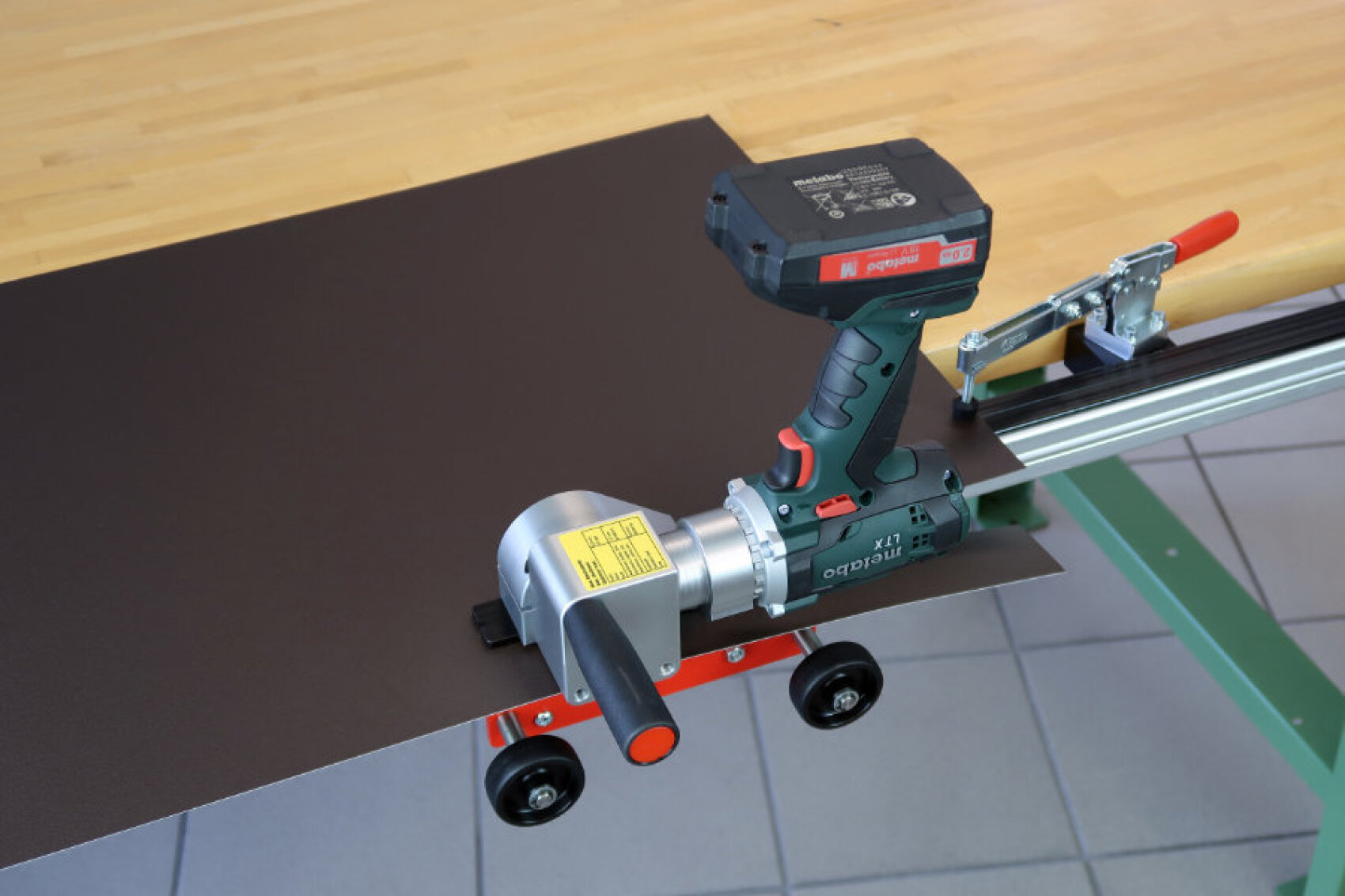 A cordless electric sheet metal cutter is positioned to cut a dark sheet on a wooden workbench, with a metal guide rail attached to the table. The floor is tiled.